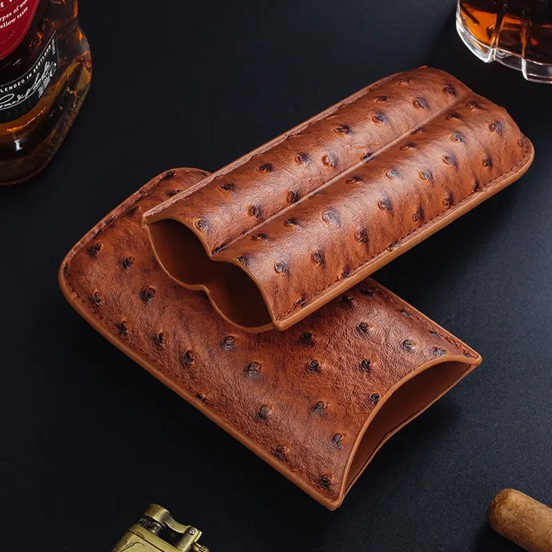 Brown leather cigar humidor on a dark surface with a bottle and glass in the background.